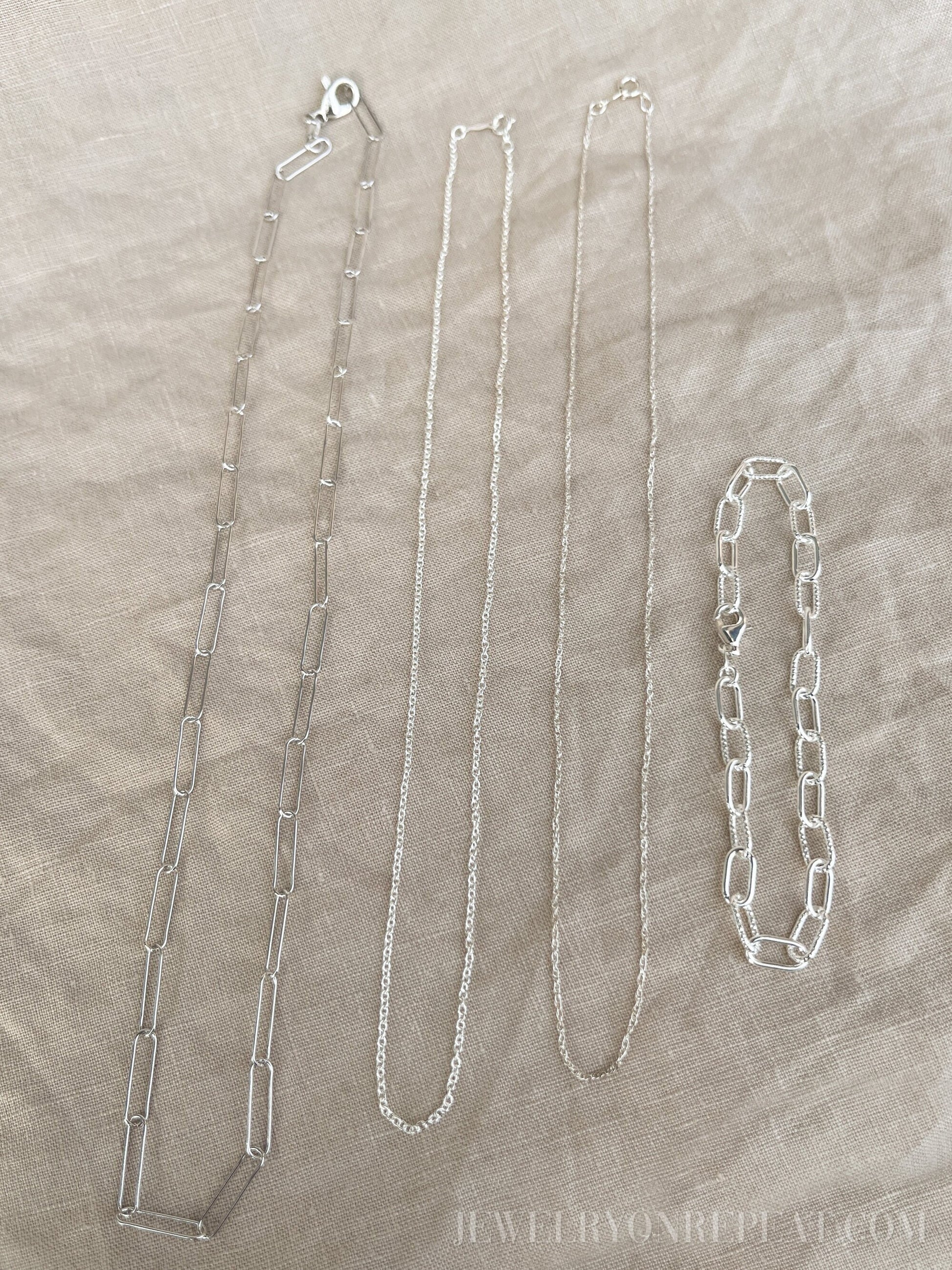 May include: Four silver chains on a beige fabric background. The chains are different lengths and styles. The chain on the left is a thick, open link chain. The chain in the middle is a thin, delicate chain. The chain on the right is a thick, closed link chain. The chain on the far right is a bracelet with a thick, closed link chain.