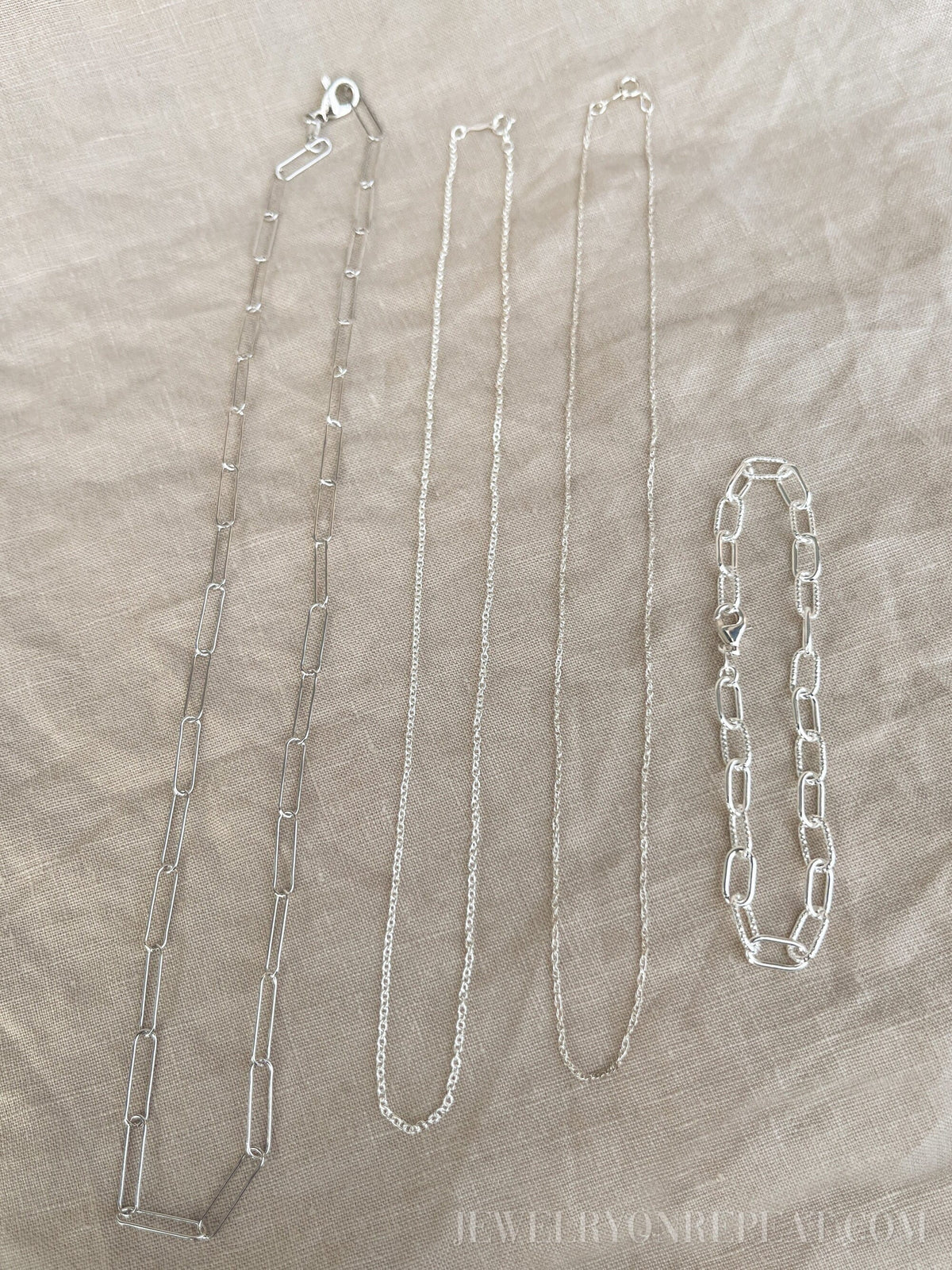 May include: Four silver chains on a beige fabric background. The chains are different lengths and styles. The chain on the left is a thick, open link chain. The chain in the middle is a thin, delicate chain. The chain on the right is a thick, closed link chain. The chain on the far right is a bracelet with a thick, closed link chain.