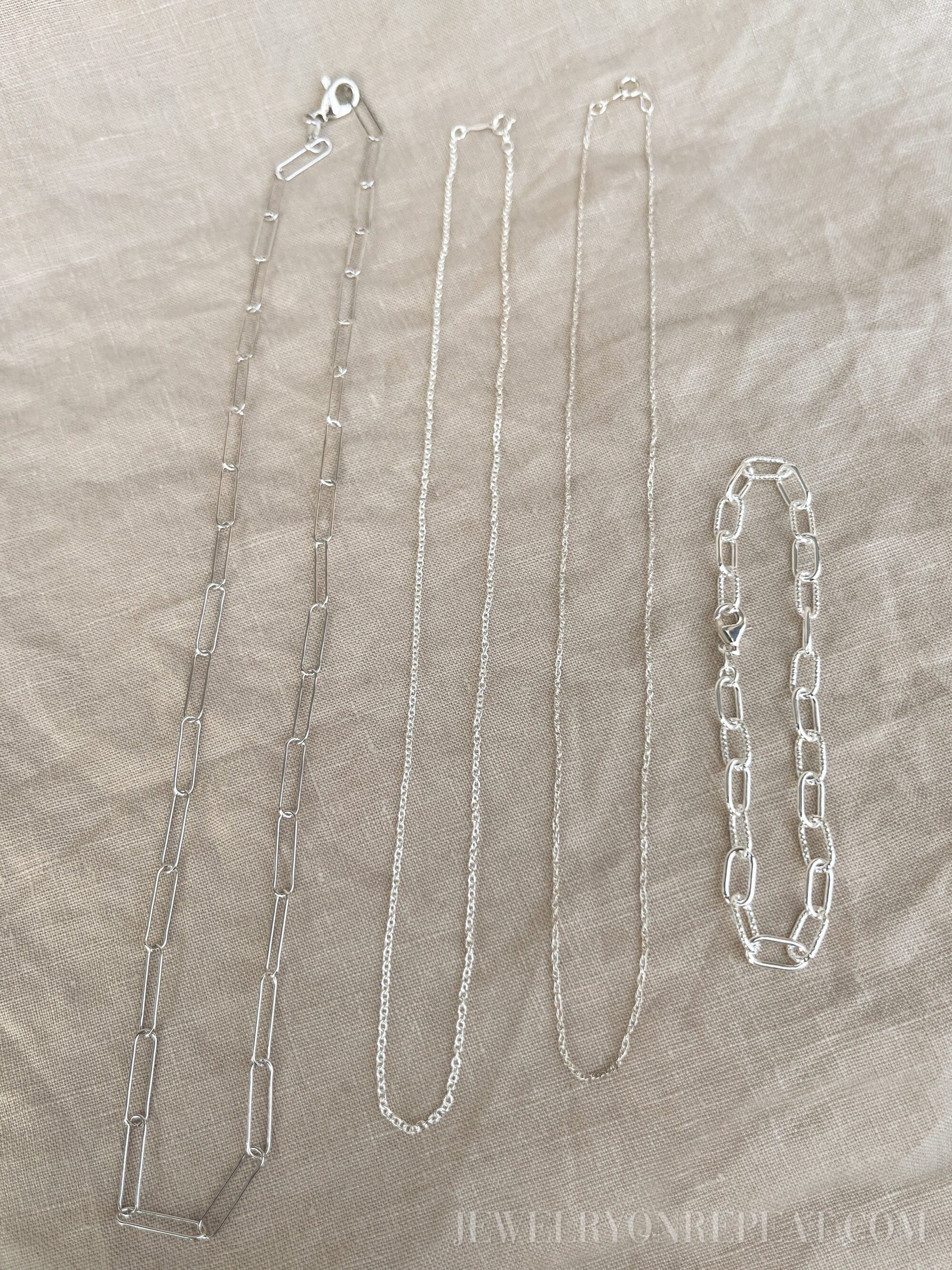 May include: Four silver chains on a beige fabric background. The chains are different lengths and styles. The chain on the left is a thick, open link chain. The chain in the middle is a thin, delicate chain. The chain on the right is a thick, closed link chain. The chain on the far right is a bracelet with a thick, closed link chain.