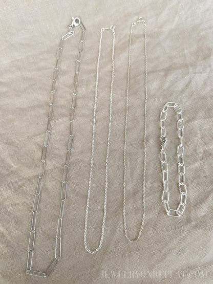 May include: Four silver chains on a beige fabric background. The chains are different lengths and styles. The chain on the left is a thick, open link chain. The chain in the middle is a thin, delicate chain. The chain on the right is a thick, closed link chain. The chain on the far right is a bracelet with a thick, closed link chain.