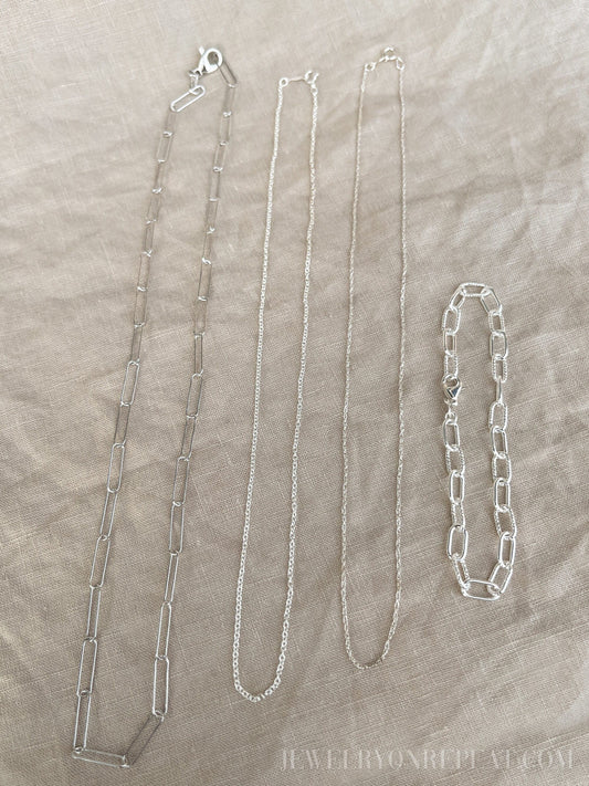 May include: Four silver chains on a beige fabric background. The chains are different lengths and styles. The chain on the left is a thick, open link chain. The chain in the middle is a thin, delicate chain. The chain on the right is a thick, closed link chain. The chain on the far right is a bracelet with a thick, closed link chain.