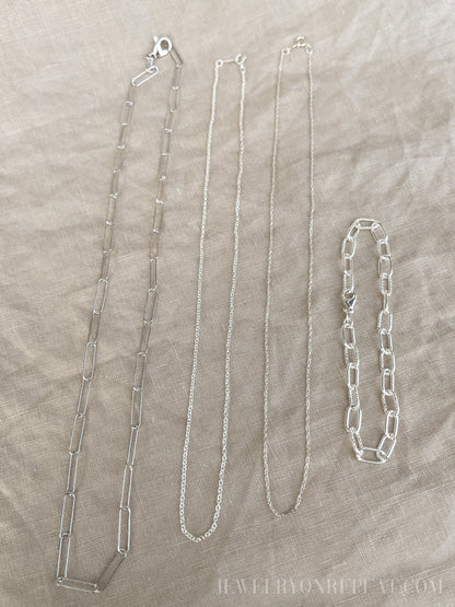 May include: Four silver chains on a beige fabric background. The chains are different lengths and styles. The chain on the left is a thick, open link chain. The chain in the middle is a thin, delicate chain. The chain on the right is a thick, closed link chain. The chain on the far right is a bracelet with a thick, closed link chain.