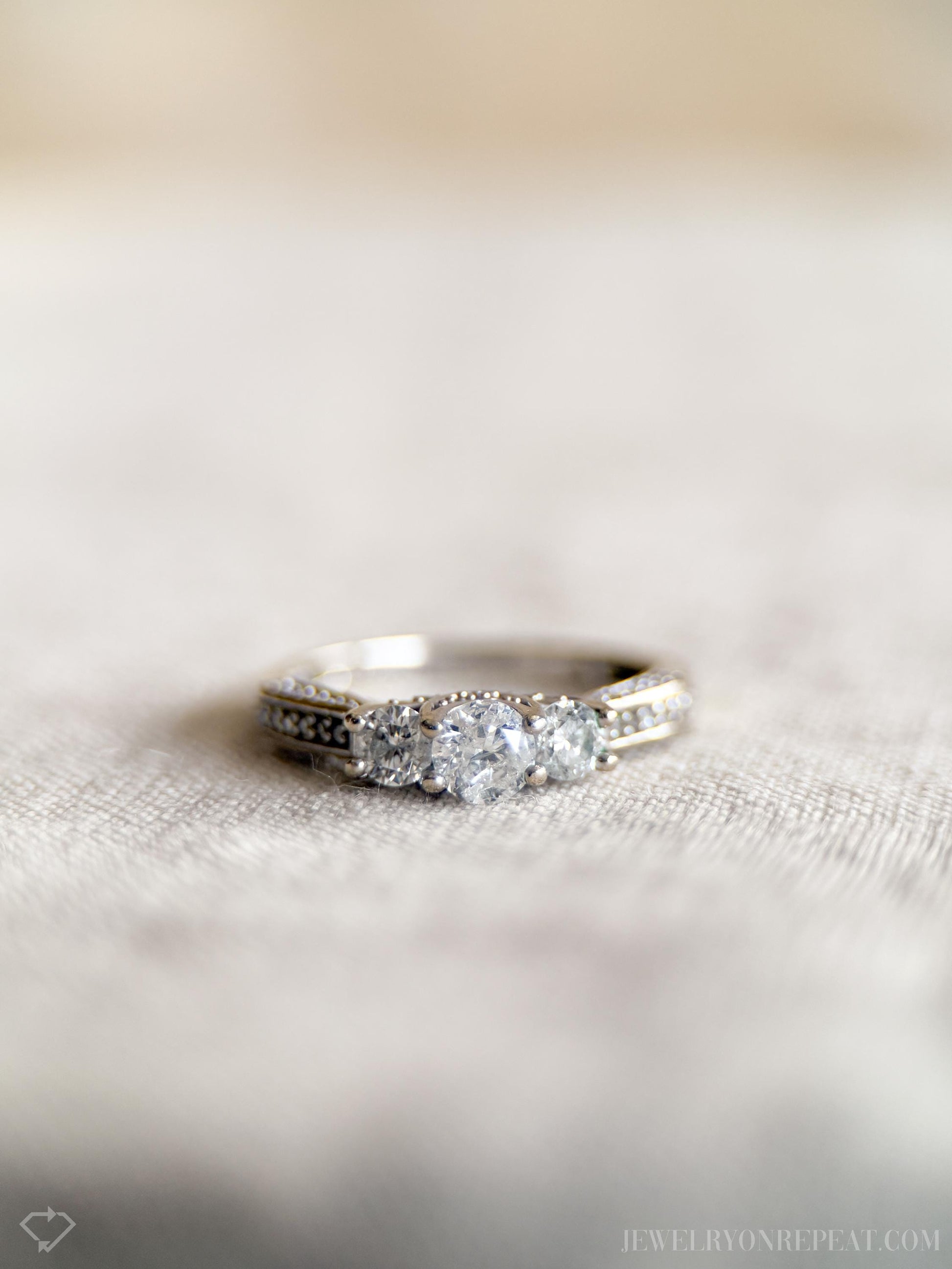 May include: Close-up of a three-stone diamond ring on a light beige fabric. The ring features a larger center diamond flanked by two smaller diamonds, all set in a white metal band with small pave diamonds along the shank. The diamonds are clear and sparkly.  The setting appears to be a classic three-stone design. Jewelry, diamond ring, three-stone, pave, engagement ring.