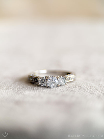May include: Close-up of a three-stone diamond ring on a light beige fabric. The ring features a larger center diamond flanked by two smaller diamonds, all set in a white metal band with small pave diamonds along the shank. The diamonds are clear and sparkly.  The setting appears to be a classic three-stone design. Jewelry, diamond ring, three-stone, pave, engagement ring.