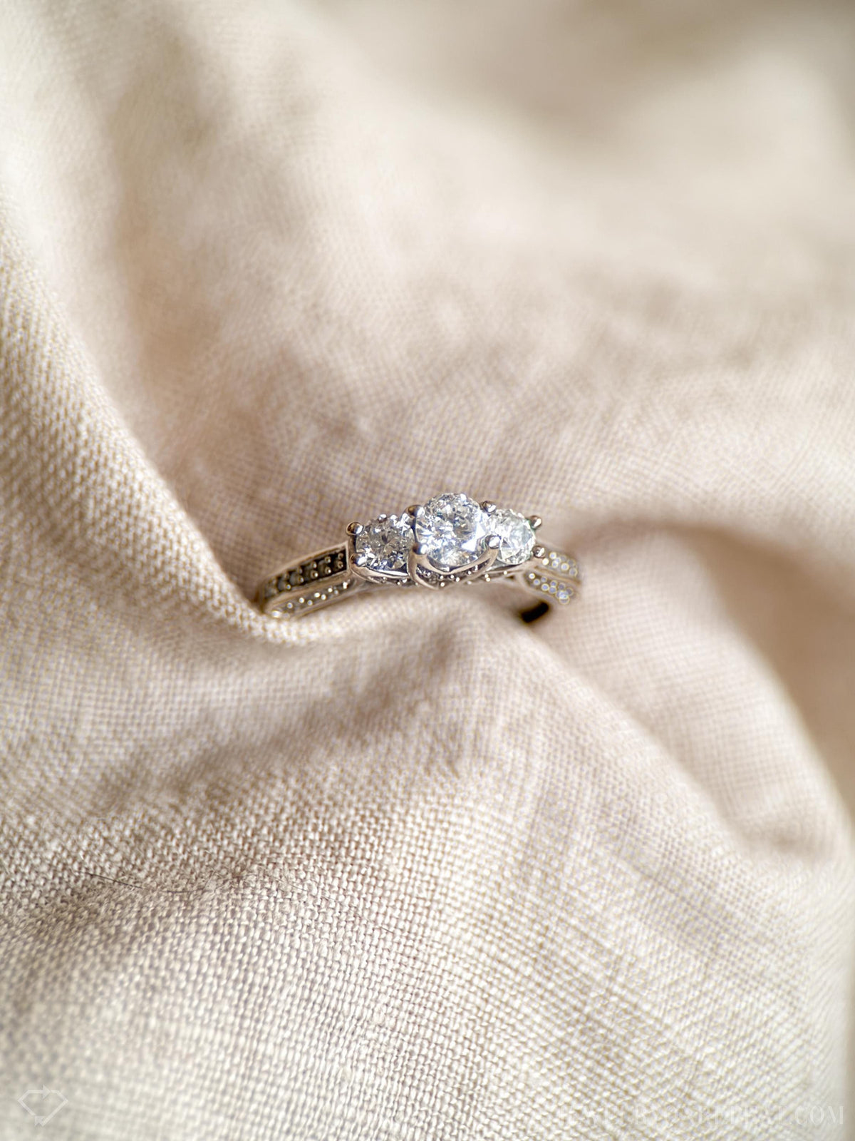May include: Close-up view of a three-stone diamond engagement ring. The ring features a central oval-shaped diamond flanked by two smaller round diamonds. The band is adorned with pave-set smaller diamonds. The ring is elegantly displayed on a light beige fabric background.