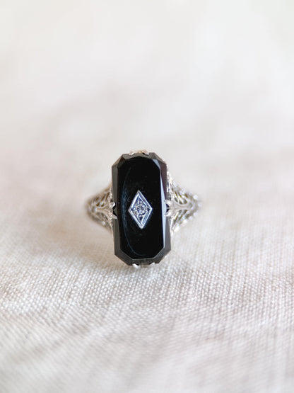 May include: An antique ring featuring a rectangular black onyx stone with a small diamond accent. The ring has an ornate, filigree design on the band, crafted in silver or white gold. The ring is set against a neutral background.