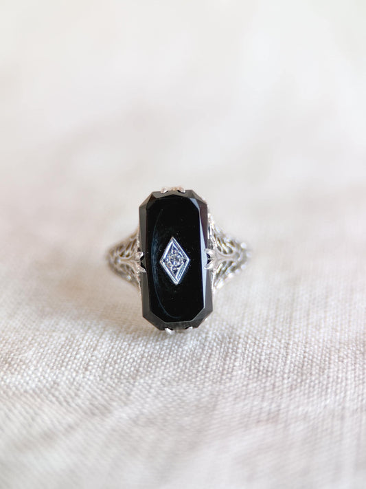 May include: An antique ring featuring a rectangular black onyx stone with a small diamond accent. The ring has an ornate, filigree design on the band, crafted in silver or white gold. The ring is set against a neutral background.