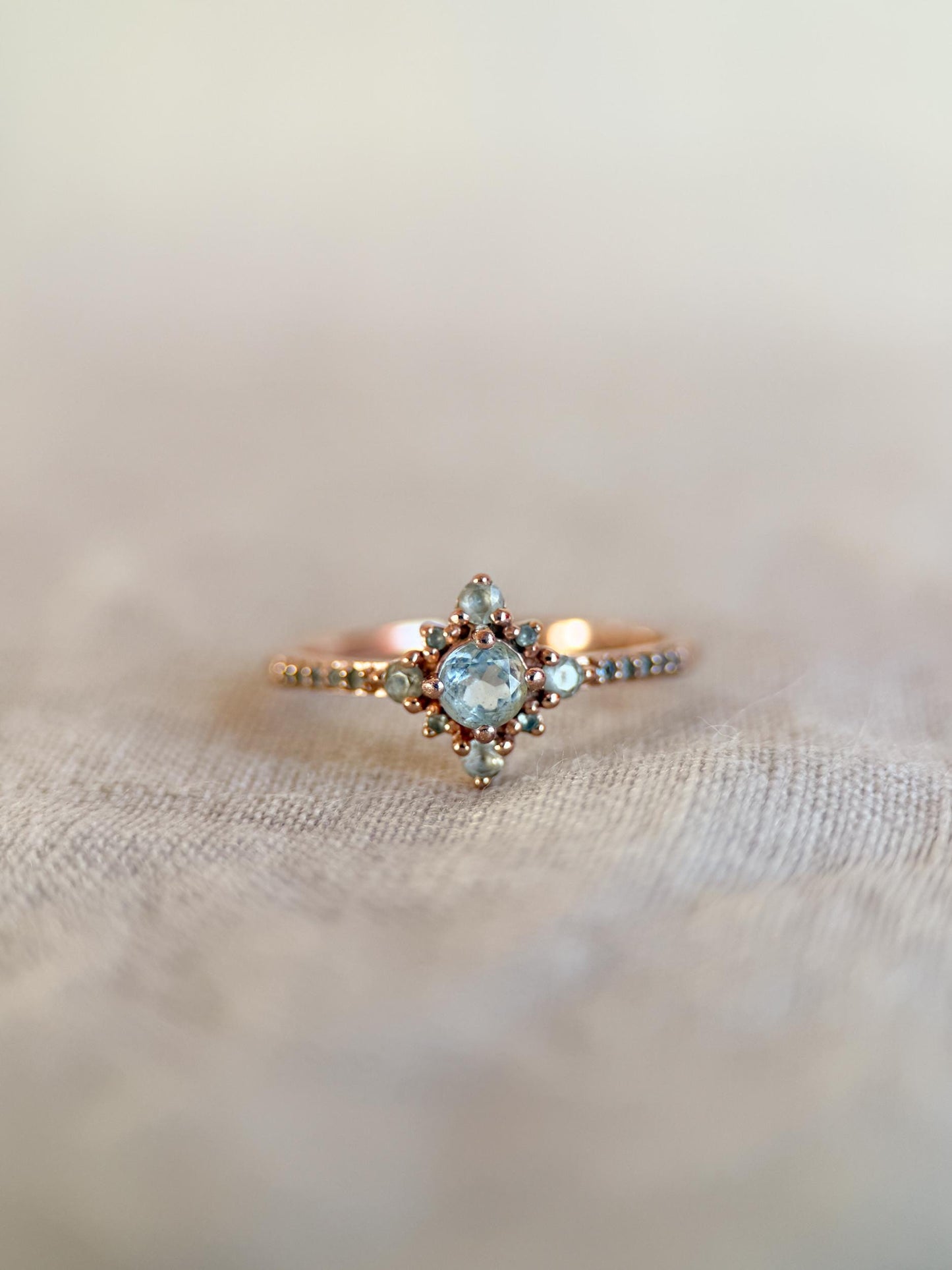 May include: A rose gold ring featuring a central light blue gemstone surrounded by smaller, matching stones in a starburst design. The band is adorned with tiny, evenly spaced beads. The ring is set against a soft, neutral background.