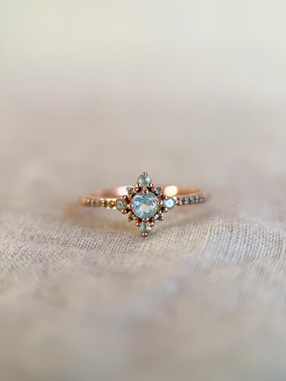 May include: A rose gold ring featuring a central light blue gemstone surrounded by smaller, matching stones in a starburst design. The band is adorned with tiny, evenly spaced beads. The ring is set against a soft, neutral background.