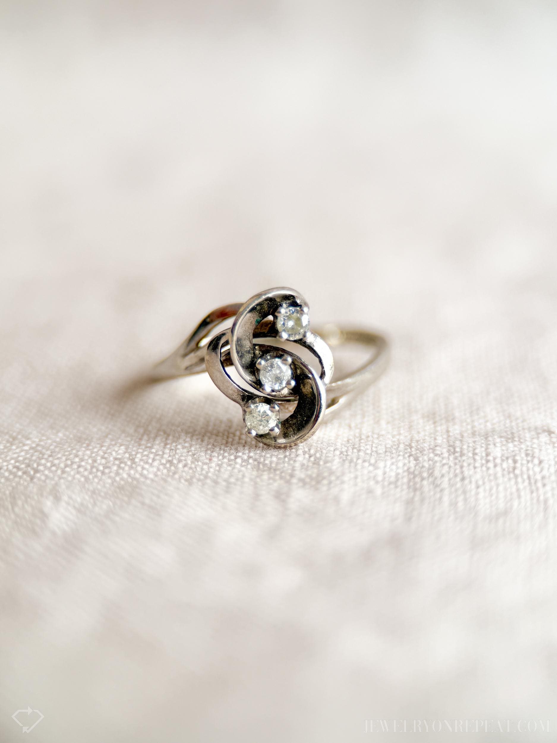 May include: A silver ring with three small, round, clear gemstones. The ring has a unique, swirling design with the gemstones set in the center. The ring is displayed on a light-colored, textured surface.
