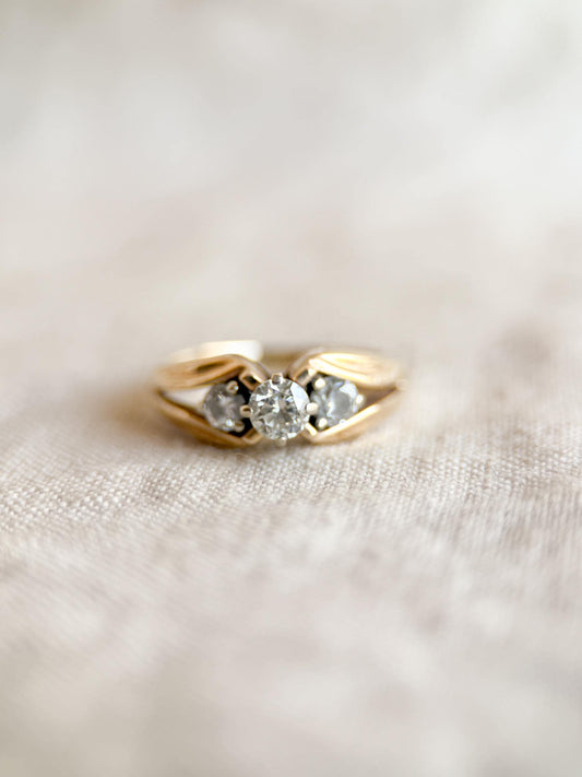 May include: A gold ring with three clear gemstones. The ring has a classic design with a band that splits to hold the gemstones. The gemstones are round and set in a prong setting. The ring is on a neutral-colored fabric.