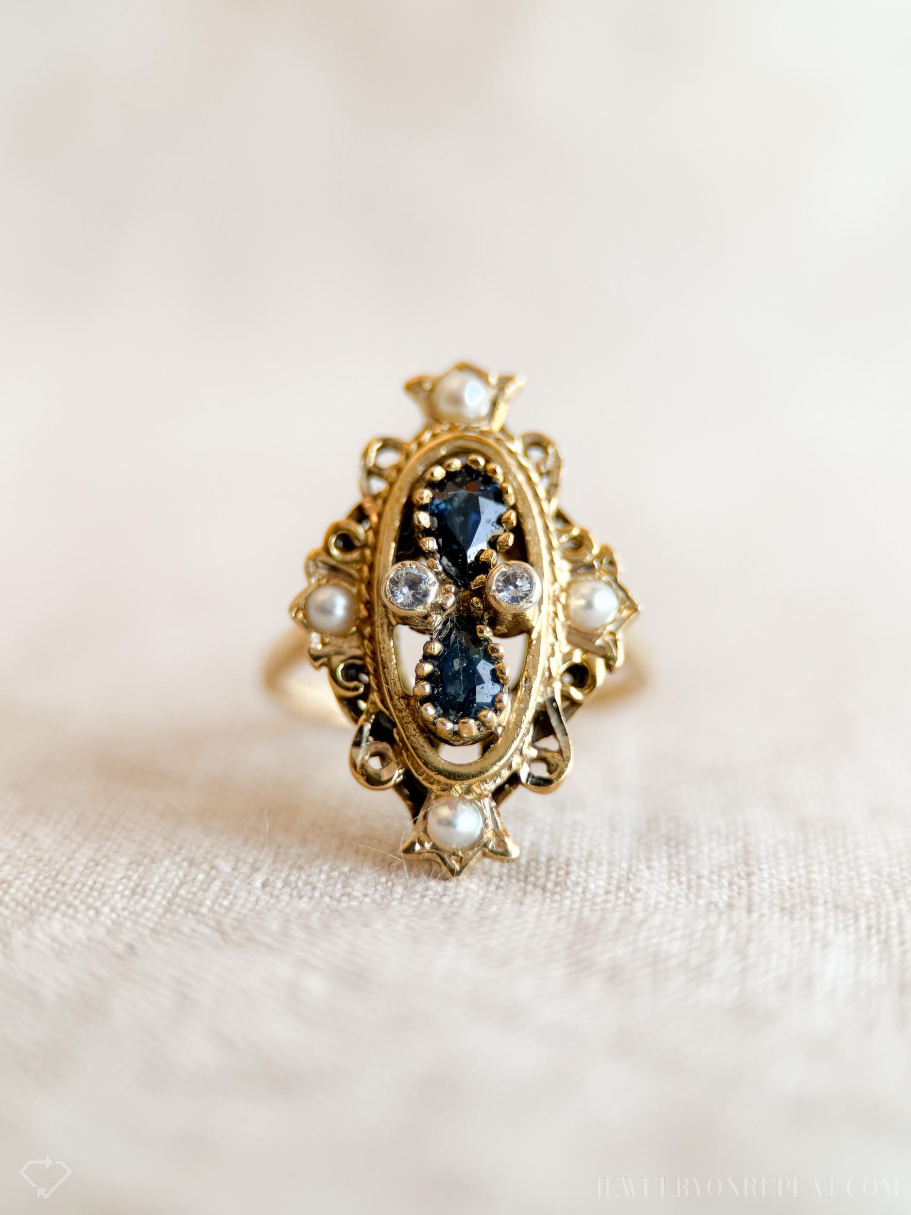 May include: A gold-toned ring with an oval centerpiece. The ring features dark blue gemstones, small clear stones, and white pearls. The ornate design includes filigree and decorative elements. The ring is set against a neutral background.