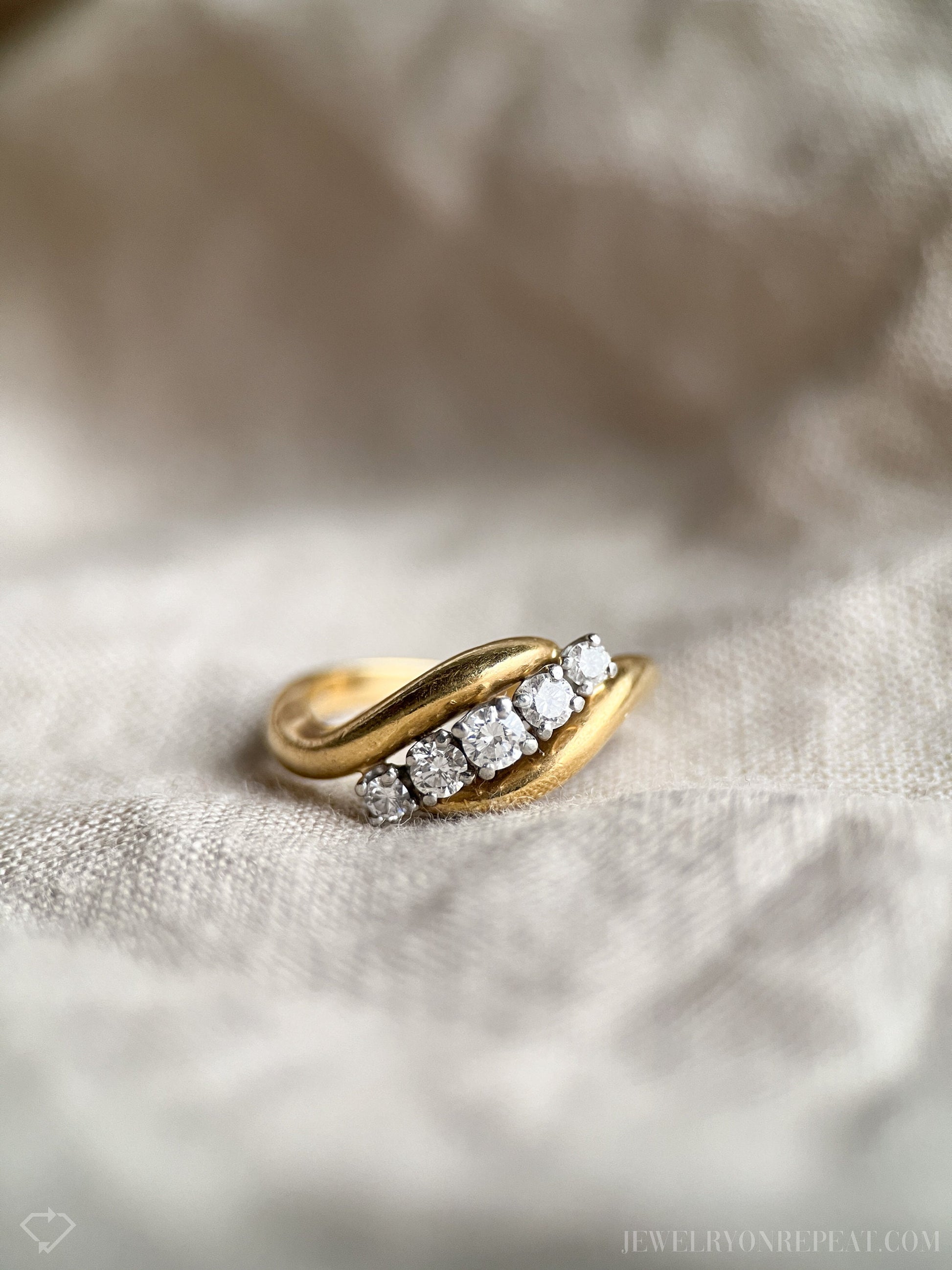 May include: A gold band ring with five round diamonds set in a curved line. The ring is on a white fabric background.