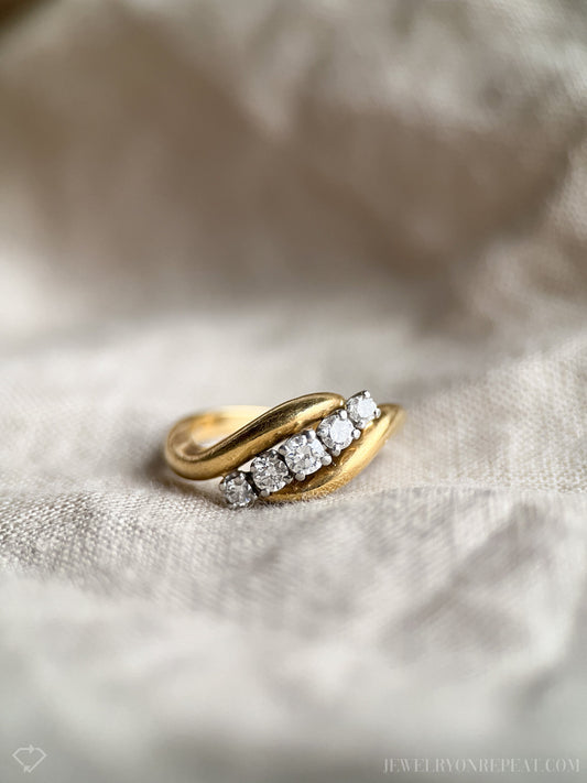 May include: A gold band ring with five round diamonds set in a curved line. The ring is on a white fabric background.