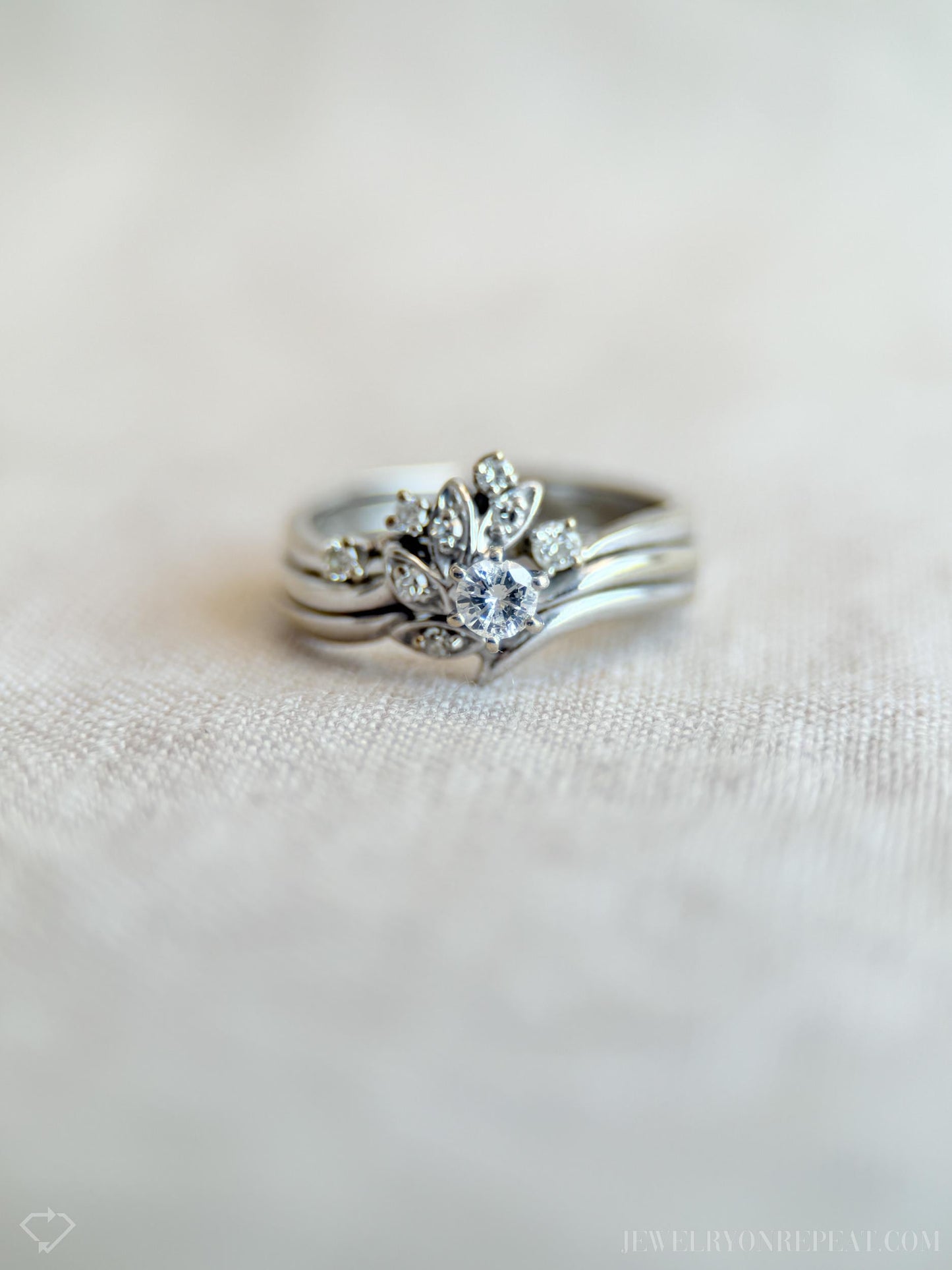 May include: A silver ring set featuring a central ring with a large, round, clear gemstone surrounded by smaller stones and leaf-like designs. The set includes two additional bands. The rings are displayed on a light-colored, textured surface.