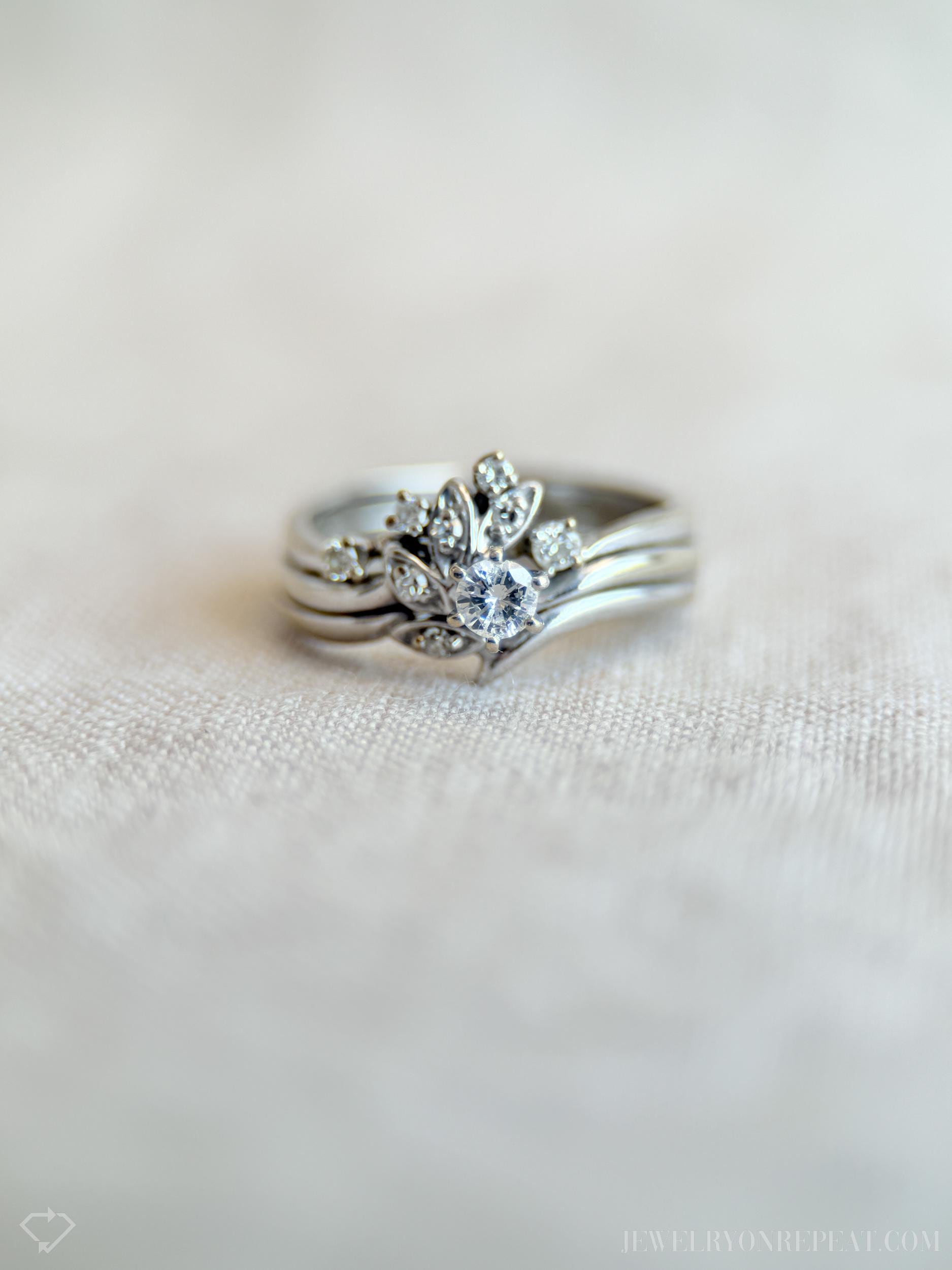 May include: A silver ring set featuring a central ring with a large, round, clear gemstone surrounded by smaller stones and leaf-like designs. The set includes two additional bands. The rings are displayed on a light-colored, textured surface.