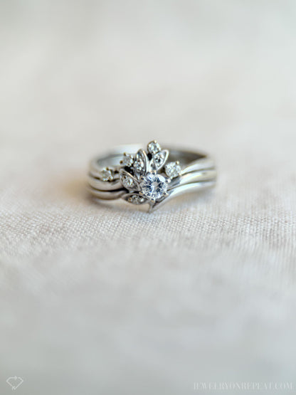 May include: A silver ring set featuring a central ring with a large, round, clear gemstone surrounded by smaller stones and leaf-like designs. The set includes two additional bands. The rings are displayed on a light-colored, textured surface.