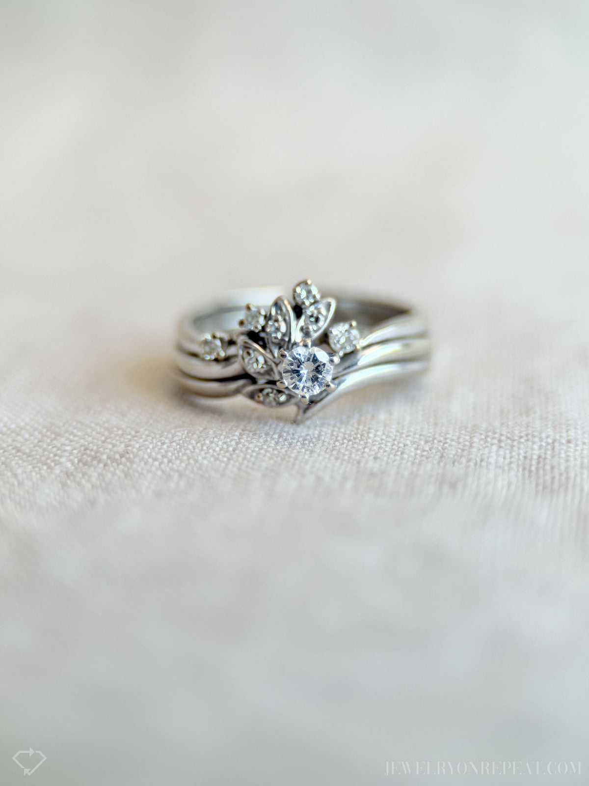 May include: A silver ring set featuring a central ring with a large, round, clear gemstone surrounded by smaller stones and leaf-like designs. The set includes two additional bands. The rings are displayed on a light-colored, textured surface.