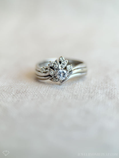 May include: A silver ring set featuring a central ring with a large, round, clear gemstone surrounded by smaller stones and leaf-like designs. The set includes two additional bands. The rings are displayed on a light-colored, textured surface.