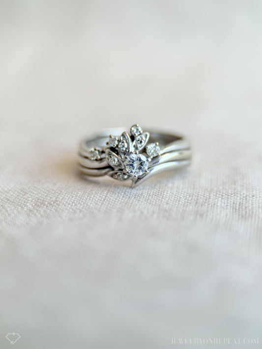 May include: A silver ring set featuring a central ring with a large, round, clear gemstone surrounded by smaller stones and leaf-like designs. The set includes two additional bands. The rings are displayed on a light-colored, textured surface.