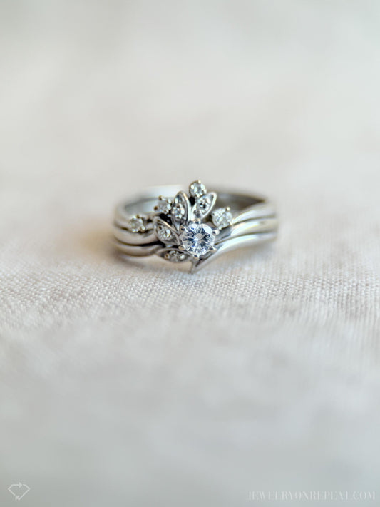 May include: A silver ring set featuring a central ring with a large, round, clear gemstone surrounded by smaller stones and leaf-like designs. The set includes two additional bands. The rings are displayed on a light-colored, textured surface.