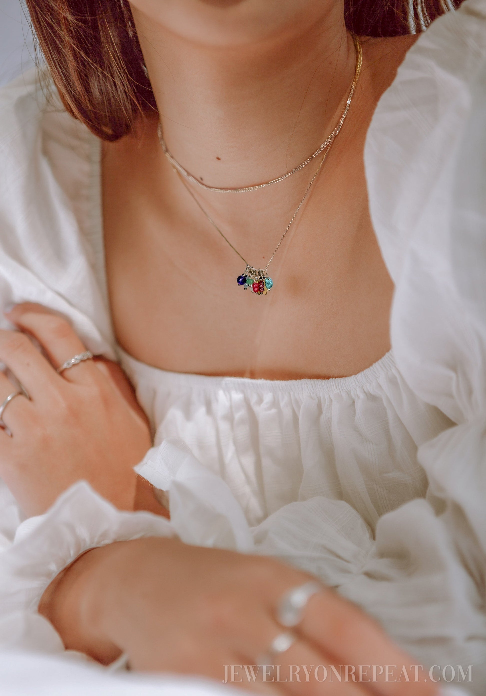 May include: A silver chain necklace with a charm featuring multiple colorful gemstones. The necklace is layered over a thin silver chain.