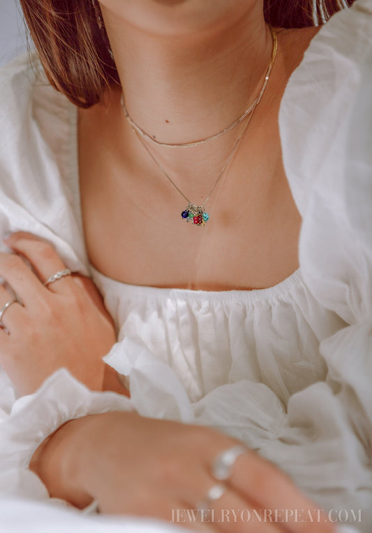 May include: A silver chain necklace with a charm featuring multiple colorful gemstones. The necklace is layered over a thin silver chain.