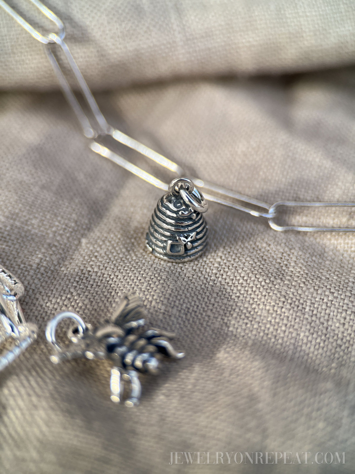 May include: Close-up of a silver-toned charm necklace with a beehive and bee pendant. The beehive charm is detailed with horizontal lines and a small bee. The bee charm is also silver-toned. The necklace is on a neutral-colored fabric.
