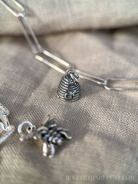May include: Close-up of a silver-toned charm necklace with a beehive and bee pendant. The beehive charm is detailed with horizontal lines and a small bee. The bee charm is also silver-toned. The necklace is on a neutral-colored fabric.
