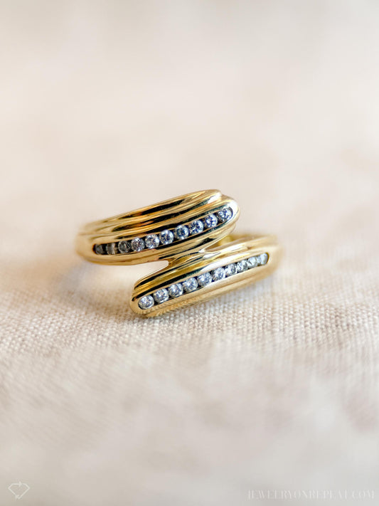 May include: A gold-colored ring with a wrap-around design. The ring features two curved bands, each adorned with a row of small, clear gemstones. The ring is set against a neutral, textured background, highlighting its elegant design.