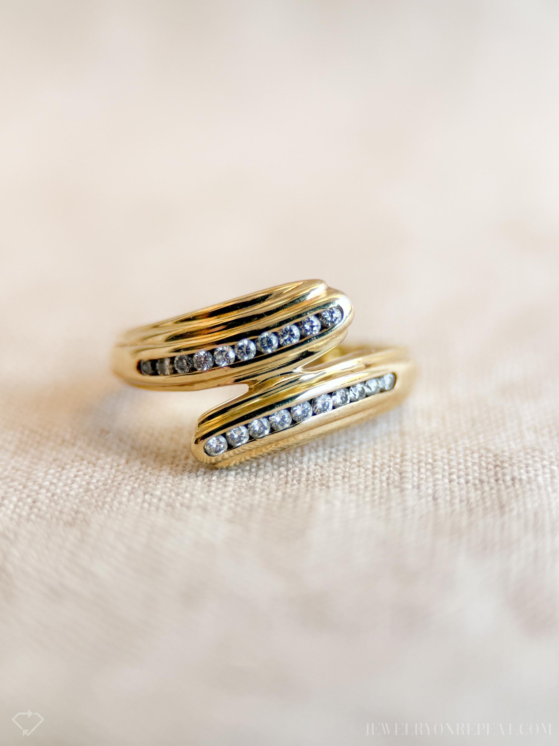 May include: A gold-colored ring with a wrap-around design. The ring features two curved bands, each adorned with a row of small, clear gemstones. The ring is set against a neutral, textured background, highlighting its elegant design.