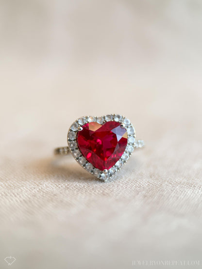 May include: A heart-shaped ring with a large, faceted red gemstone at the center. The gemstone is surrounded by a halo of smaller, clear stones set in a silver band. The ring is displayed on a neutral-toned surface.