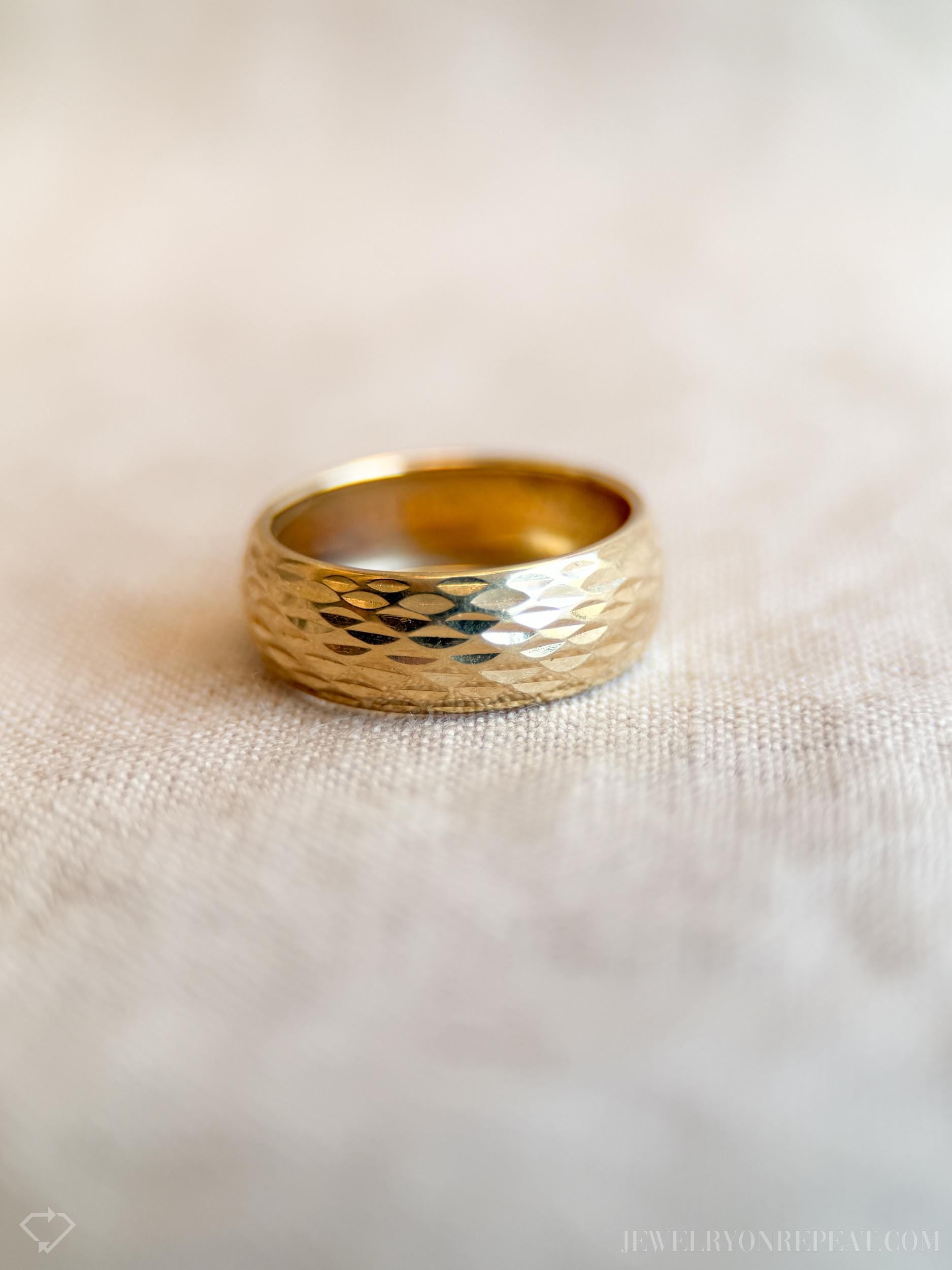 May include: A gold ring with a textured, hammered design. The wide band ring has a shiny, reflective surface and is set against a neutral background. The ring's surface is covered in small, geometric facets.