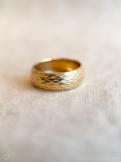 May include: A gold ring with a textured, hammered design. The wide band ring has a shiny, reflective surface and is set against a neutral background. The ring's surface is covered in small, geometric facets.