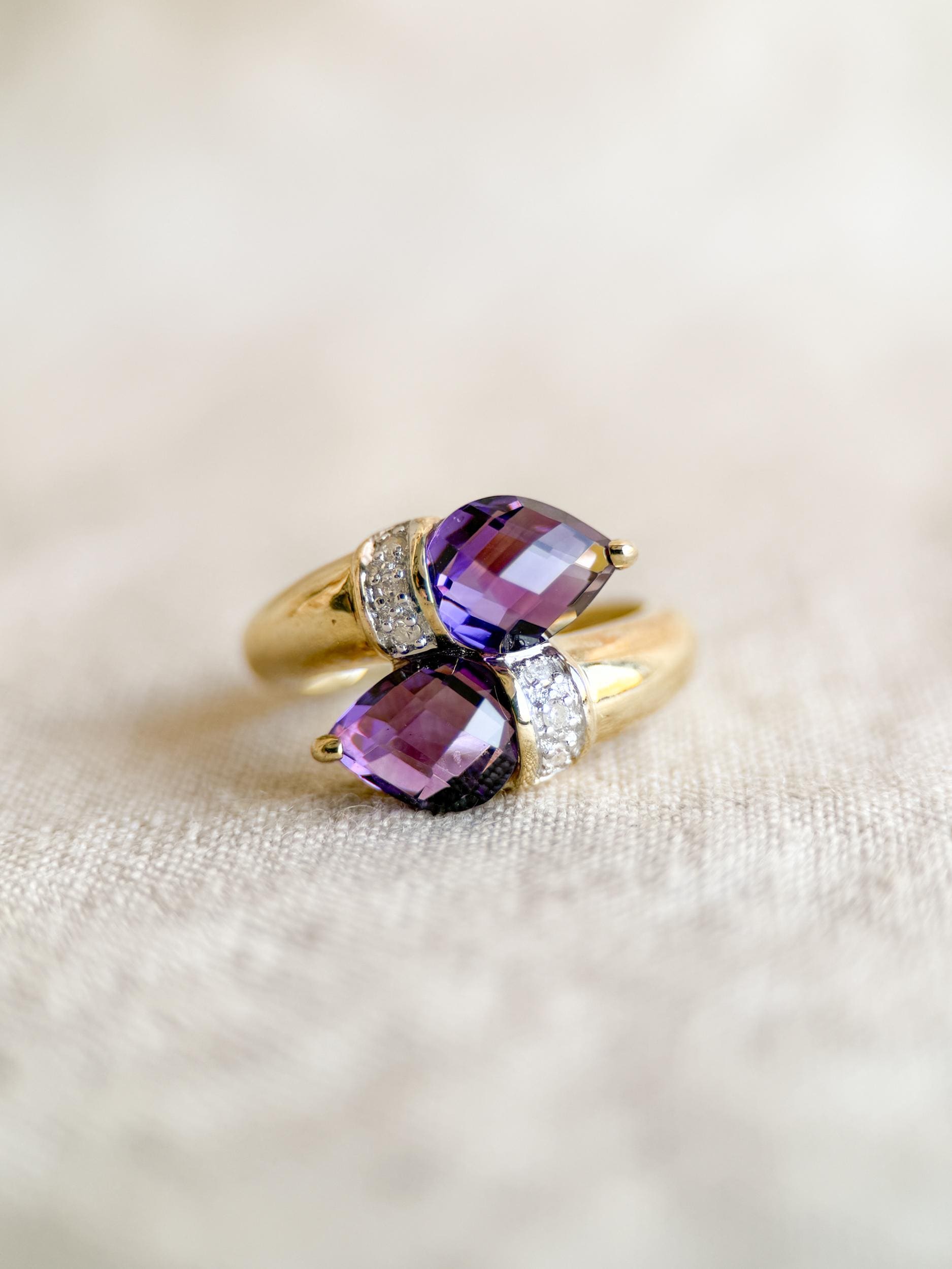 May include: A gold ring featuring two pear-shaped, deep purple gemstones. The ring's band curves around the stones, with small, clear accent stones set on each side of the purple gems. The ring is set against a neutral background.