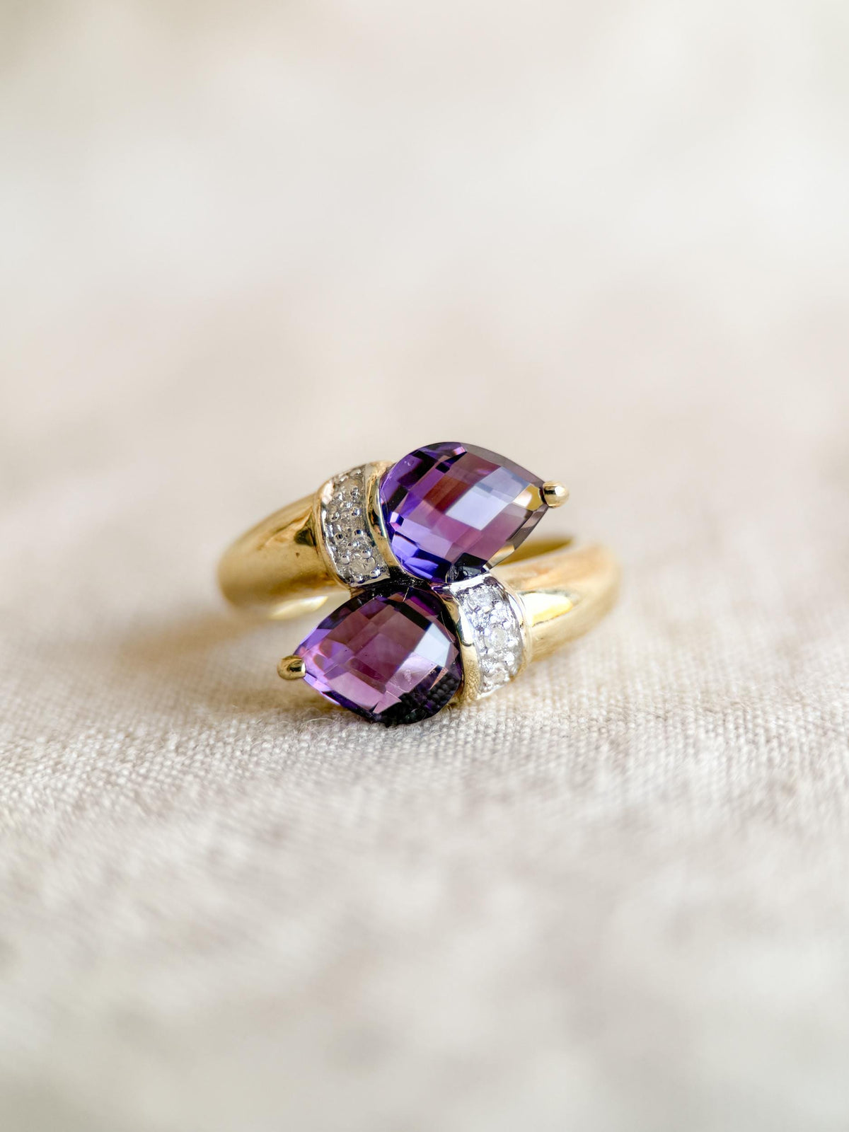 May include: A gold ring featuring two pear-shaped, deep purple gemstones. The ring's band curves around the stones, with small, clear accent stones set on each side of the purple gems. The ring is set against a neutral background.