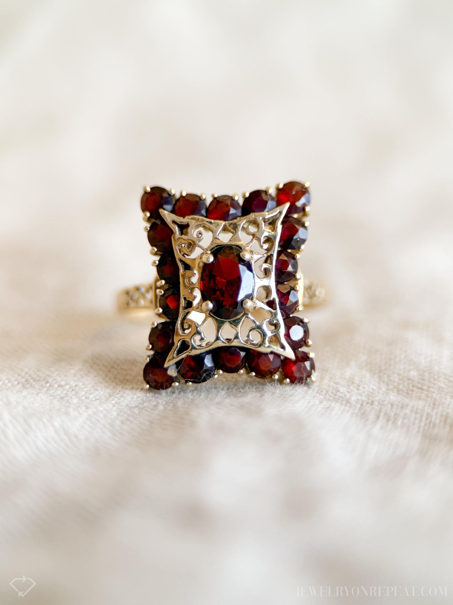 May include: A vintage gold ring featuring a square cluster of deep red gemstones. The center stone is surrounded by smaller gems and ornate filigree. The ring's band has a textured design, showcasing a classic and elegant style.