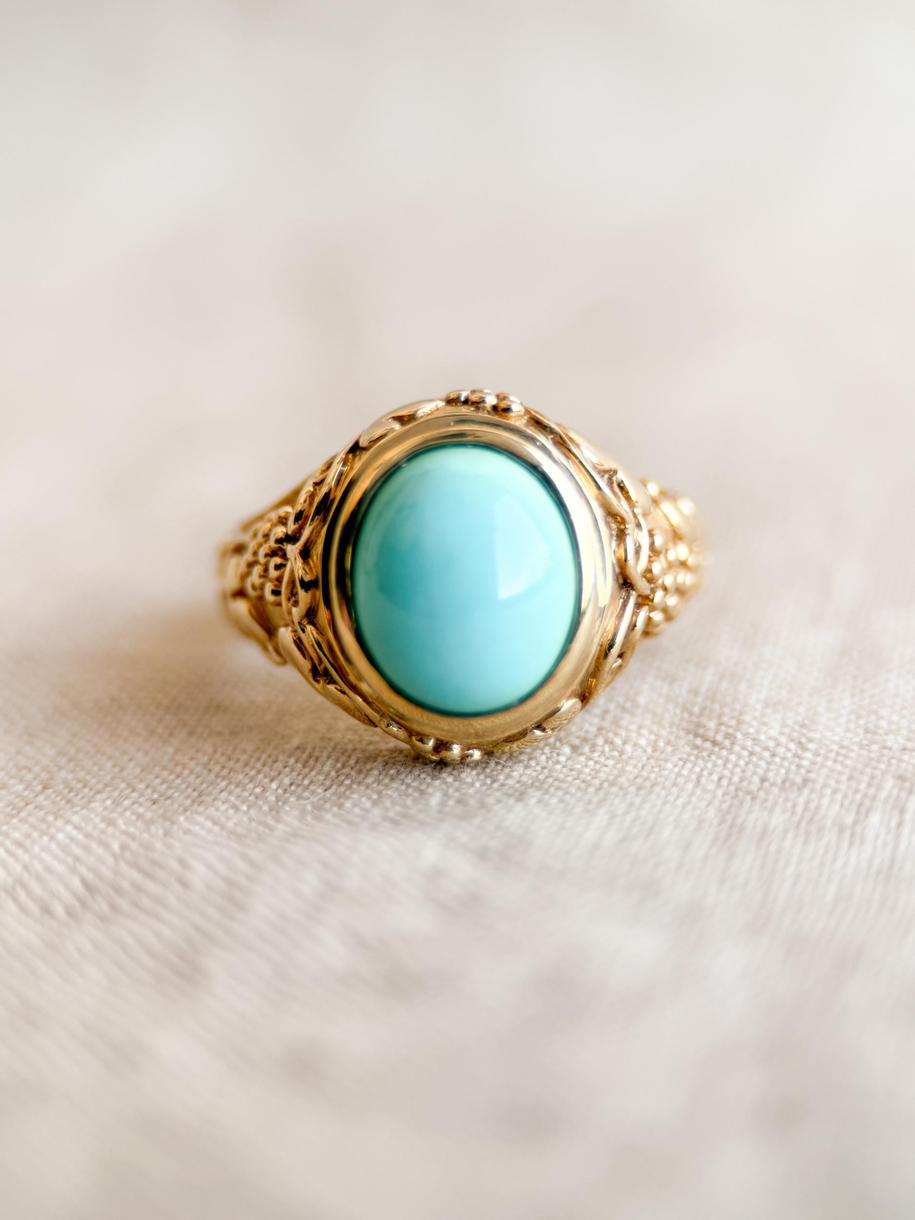 May include: A gold-colored ring with a large, oval, light blue stone. The ring has an ornate, textured band with a detailed design around the stone. The ring is set against a neutral background.