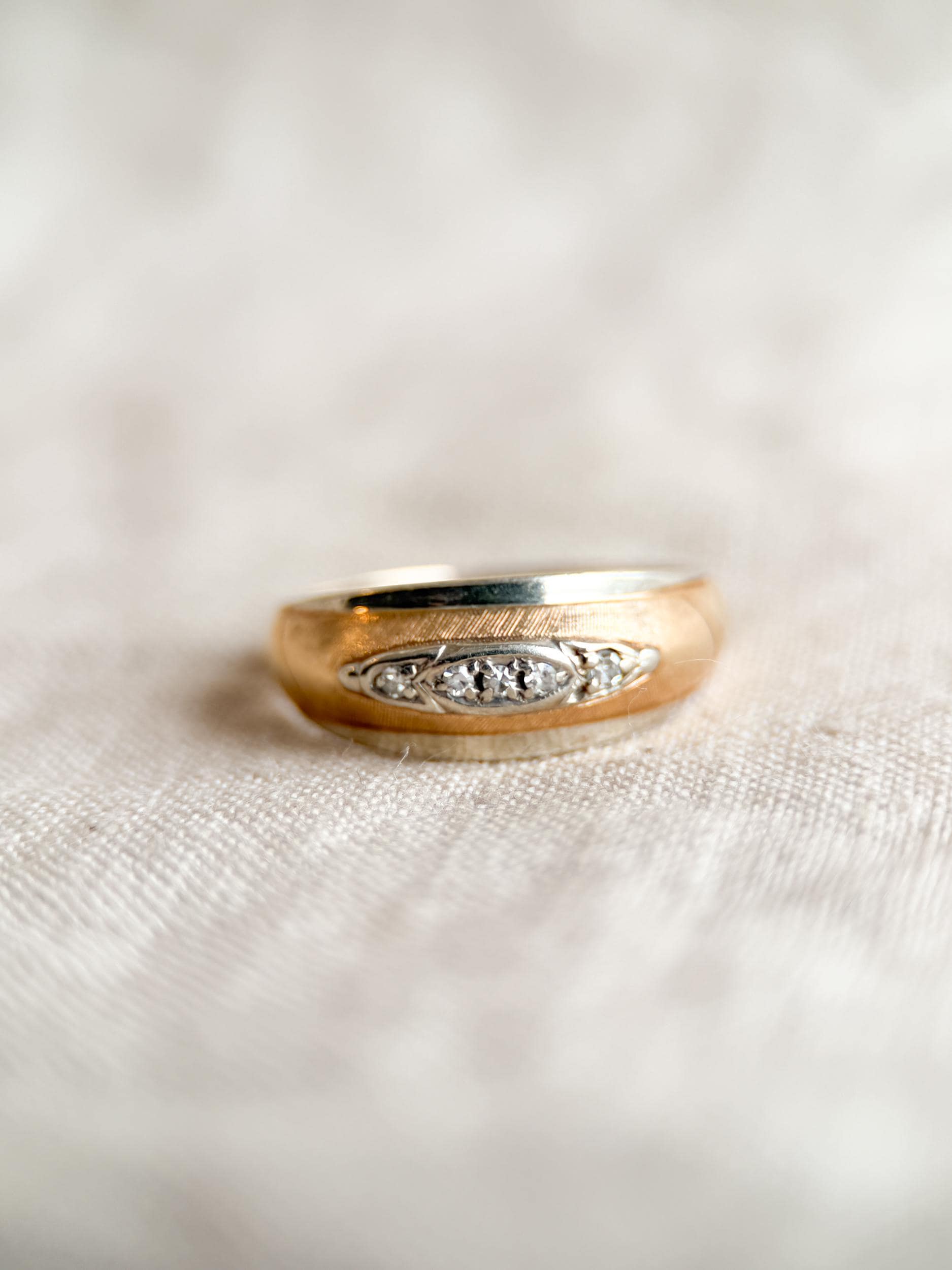 May include: A gold-toned ring with a brushed finish and a band of small, clear gemstones. The ring is set against a neutral, textured background, highlighting its classic design and elegant details. The ring's design features a central row of gemstones.