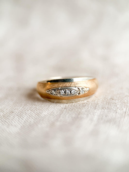 May include: A gold-toned ring with a brushed finish and a band of small, clear gemstones. The ring is set against a neutral, textured background, highlighting its classic design and elegant details. The ring's design features a central row of gemstones.