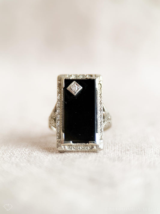 May include: An antique rectangular ring with a black onyx center stone and a small diamond accent. The ring has an ornate, filigree-style setting in a silver tone, showcasing a vintage aesthetic.