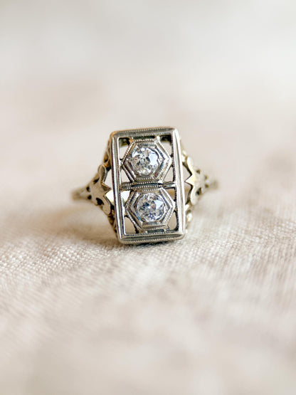 May include: A vintage-style ring with a rectangular face, featuring two hexagonal-shaped diamonds set within a filigree design. The ring has an ornate, detailed band and a silver or white gold finish, showcasing a classic, elegant aesthetic.