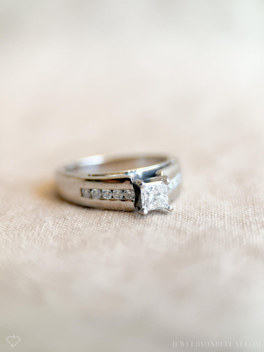 May include: A silver engagement ring with a square-cut diamond center stone. The band features a channel setting with smaller diamonds. The ring is set on a neutral-toned surface, showcasing its elegant design. The ring is a classic piece of jewelry.