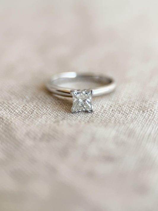May include: A silver ring with a square-cut diamond center stone. The ring band is smooth and polished, and the diamond is set in a four-prong setting. The ring is displayed on a neutral-colored fabric.