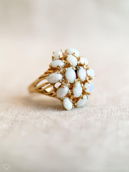 May include: A gold ring featuring a cluster of oval-shaped opals. The opals are white with a subtle iridescence, set in a detailed gold band. The ring's design is reminiscent of a floral arrangement, showcasing a vintage aesthetic.